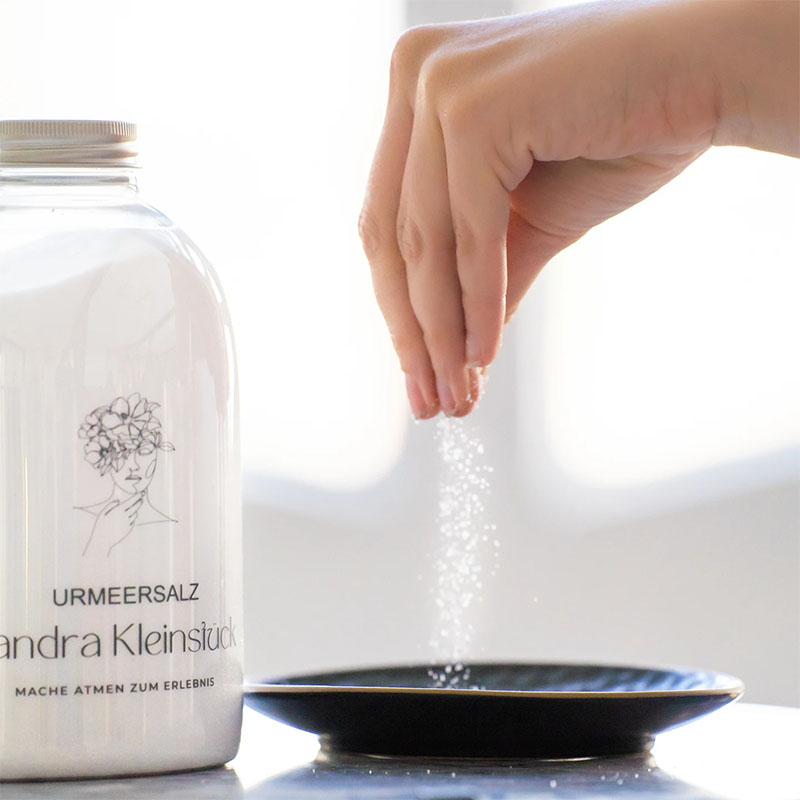 Feines Urmeersalz in Pulverform, Flasche mit feinem Urmeersalz, Hand streut dieses auf einen Teller